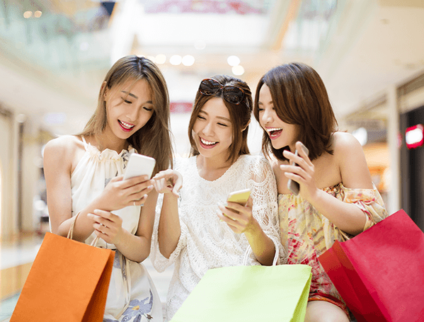 Três mulheres rindo com algumas compras em seus braços, verificando seus smartphones