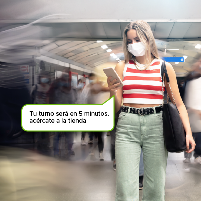 Mujer joven caminando en un centro comercial  con tapabocas mientras ve su teléfono que le notifica que su turno será pronto 