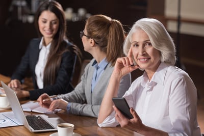 Tres mujeres en una reunión sentadas frente a sus computadoras con libretas, cafés, lápices y celulares a mano