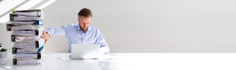 Man in blue shirt pushing away binders while using his laptop. 