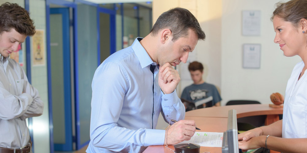 Homem de pé lendo um documento a ser assinado na frente de uma mulher em um balcão de atendimento ao cliente