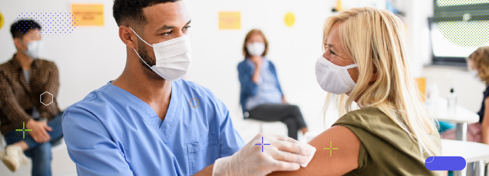 Médico hombre limpiando brazo de paciente mujer para vacunar, al fondo se ven personas esperando, todos llevan mascarilla - ACF