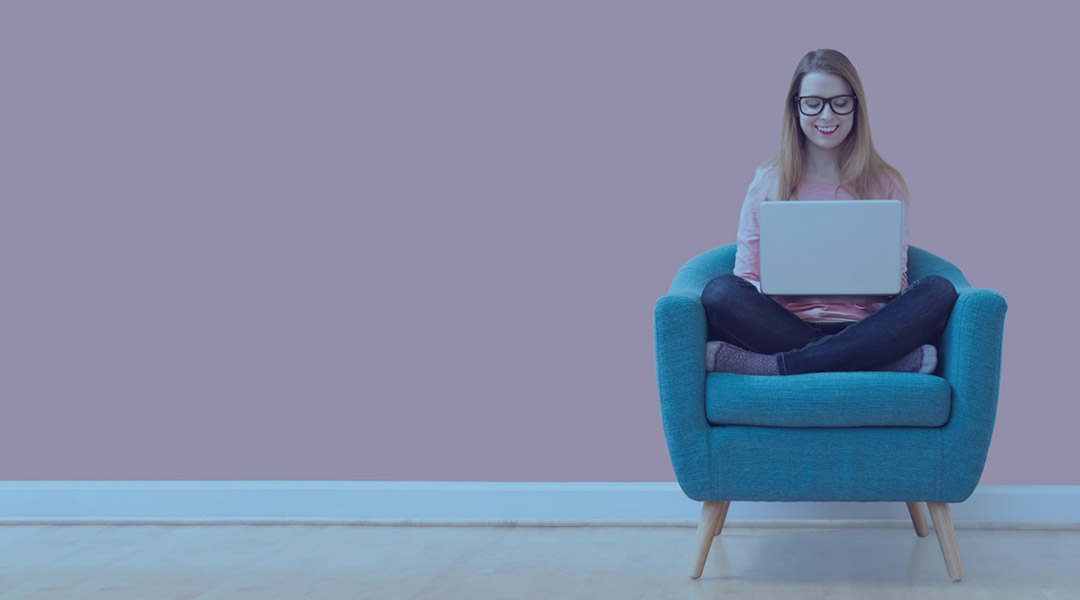 Mujer sentada sola revisando su laptop sobre un sofá de color turquesa y un fondo rosa