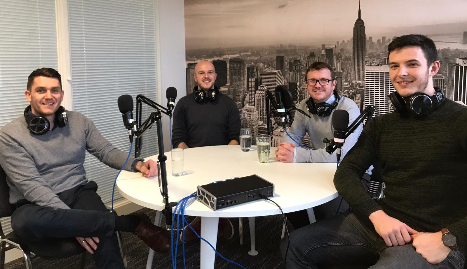 Four men sit around desk with microphones recording podcast