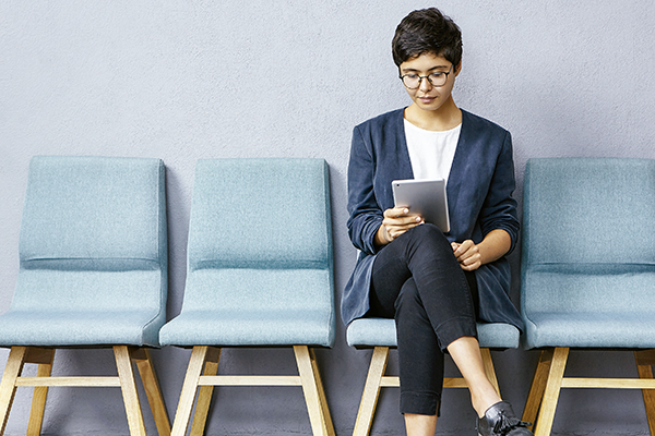 Mujer con lentes, tenida azul y negro sentada esperando ser atendida mirando su tablet