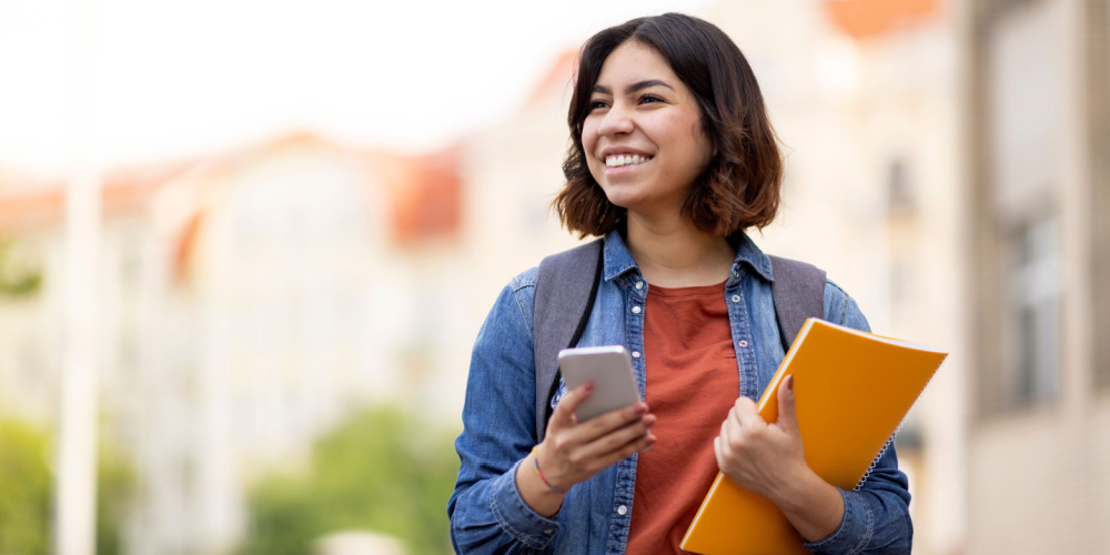 Girl carrying books and looking at her phone on a college campus.