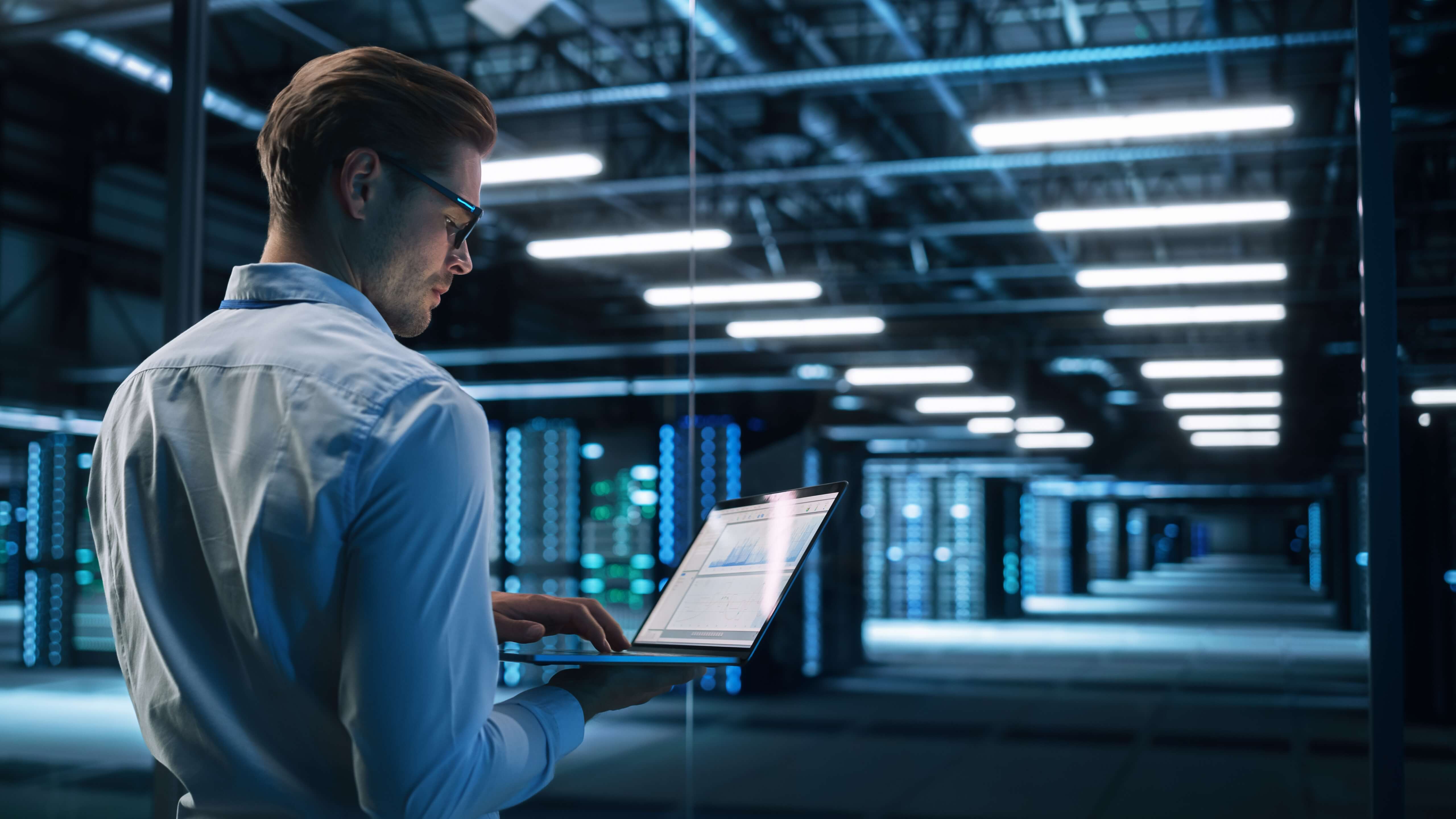 Man in blue shirt and glasses holding laptop in warehouse. 